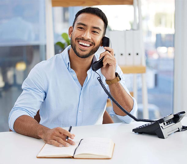 Shot of a young businessman writing in a notebook while using the phone at work