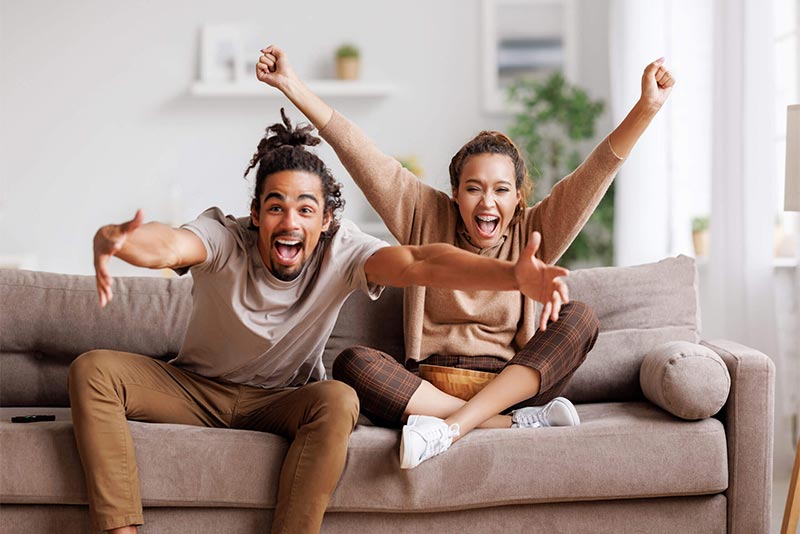 Overjoyed happy young african american couple man and woman raising arms, celebrating goal and screaming while watching baseball match together at home, selective focus. Leisure weekend activities