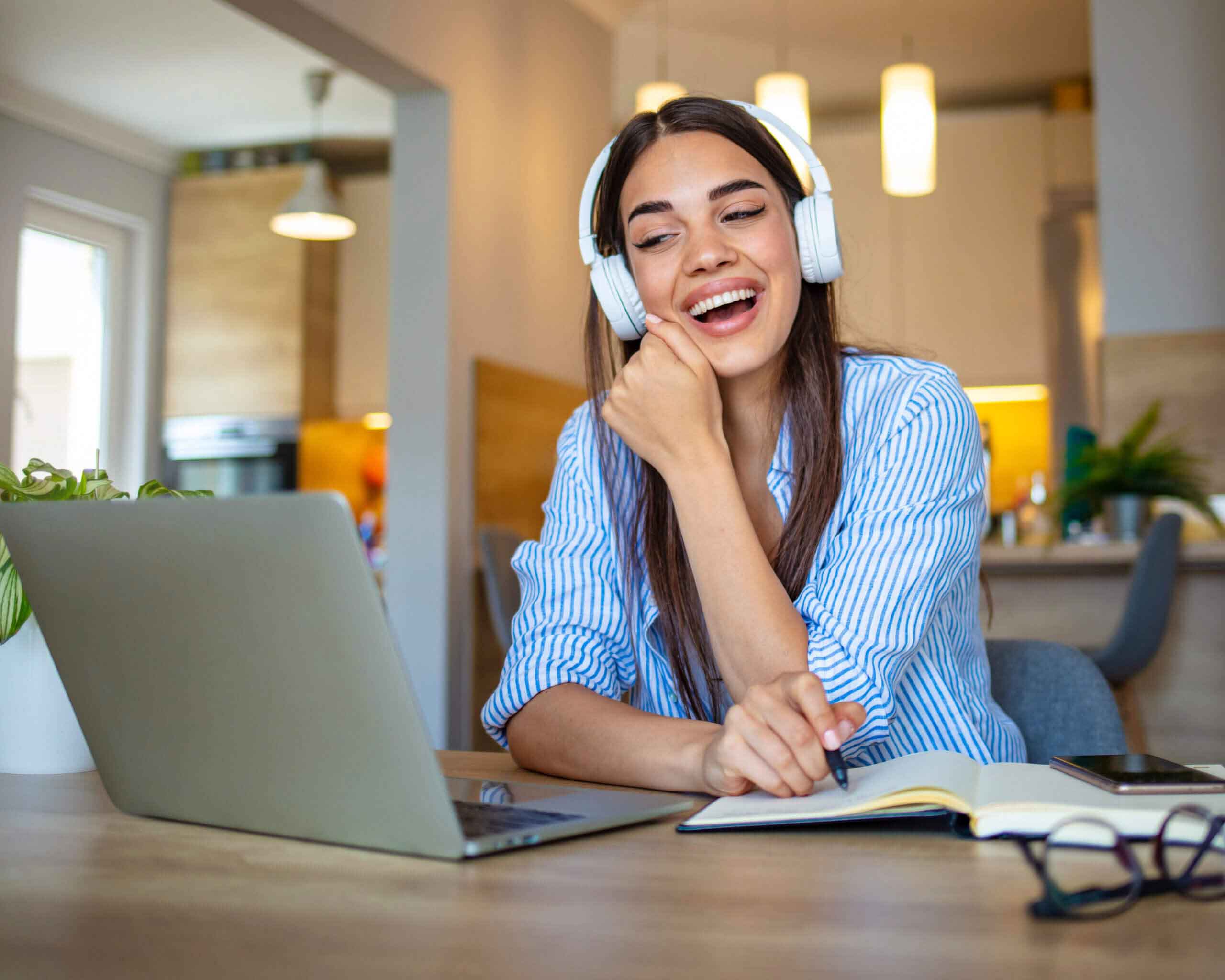 Focused woman wearing headphones using laptop at home, writing notes, attractive female student learning language, watching online webinar, listening audio course, e-learning education concept